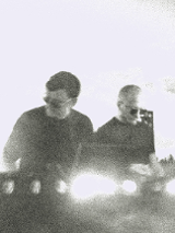 Czarno-białe, niewyraźne zdjęcie dwóch mężczyzn. Są ubrani na czarno, mają okulary przeciwsłoneczne i stoją przy konsoli DJ'a.