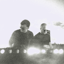 Czarno-białe, niewyraźne zdjęcie dwóch mężczyzn. Są ubrani na czarno, mają okulary przeciwsłoneczne i stoją przy konsoli DJ'a.