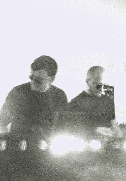 Czarno-białe, niewyraźne zdjęcie dwóch mężczyzn. Są ubrani na czarno, mają okulary przeciwsłoneczne i stoją przy konsoli DJ'a.