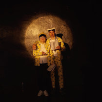 Zdjęcie z przedstawienia - kobieta i mężczyzna stoją obok siebie w zółtych strojach i czapkach na głowach, każde z nich trzyma w dłoni otwarty zeszyt.