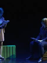 Zdjęce dwóch osób na scenie czytających tekst, jedna osoba stoi, druga osoba siedzi na skrzynce.