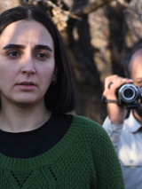 Młoda kobieta z ciemnymi włosami do ramion. Ubrana w czarną koszulkę i zielony sweter. Patrzy przejęta w dal. Za nią starszy mężczyzna z ciemnymi włosami i zarostem. Ubrany w jasną koszulę. W ręce trzyma kamerę cyfrową.