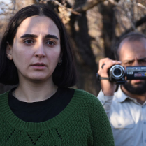 Młoda kobieta z ciemnymi włosami do ramion. Ubrana w czarną koszulkę i zielony sweter. Patrzy przejęta w dal. Za nią starszy mężczyzna z ciemnymi włosami i zarostem. Ubrany w jasną koszulę. W ręce trzyma kamerę cyfrową.