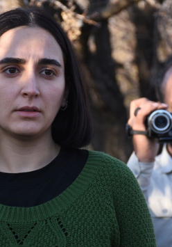 Młoda kobieta z ciemnymi włosami do ramion. Ubrana w czarną koszulkę i zielony sweter. Patrzy przejęta w dal. Za nią starszy mężczyzna z ciemnymi włosami i zarostem. Ubrany w jasną koszulę. W ręce trzyma kamerę cyfrową.