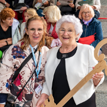Zdjęcie przedstawia szczęśliwą emerytkę trzymającą metrowy klucz do Poznania. W tle siedzą inni emeryci.