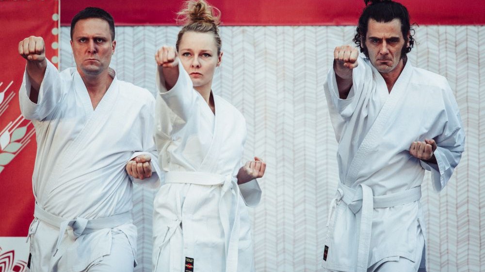 Trzy postacie ćwiczące karate, wszystkie w białych kostiumach. W środku blondynka z włosami spiętymi w niedbały kok.