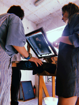 Dwie dziewczyny stoją przy maszynie do tworzenia sitodruku. Odbijają wzór na koszulce i rozmawiają przy tym.