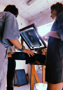 Dwie dziewczyny stoją przy maszynie do tworzenia sitodruku. Odbijają wzór na koszulce i rozmawiają przy tym.