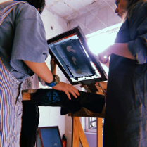 Dwie dziewczyny stoją przy maszynie do tworzenia sitodruku. Odbijają wzór na koszulce i rozmawiają przy tym.