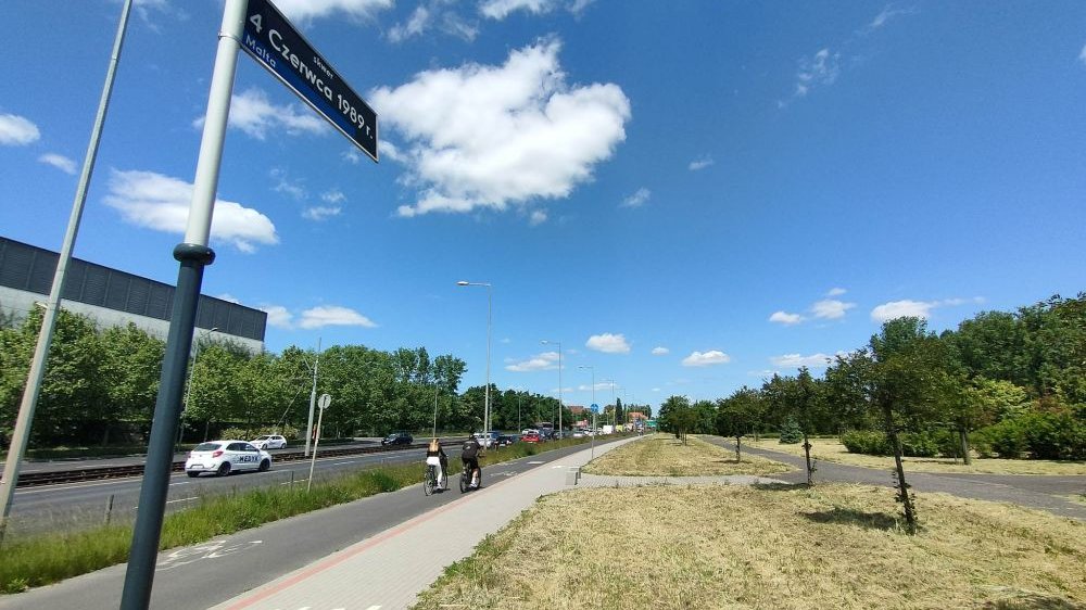 Z przodu znak drogowy z nazwą skweru. Z tyłu ulica, trawa, drzewa i ścieżka rowerowa, a na niej dwóch rowerzystów.