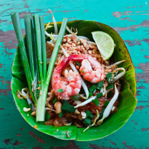 To zbliżenie talerza z danie Pad Thai, popularnym tajskim daniem z makaronem. Makaron jest smażony na wok z krewetkami, kiełkami fasoli, orzeszkami ziemnymi i szczypiorkiem. Danie podane jest na liściu banana, co jest typowe dla kuchni tajskiej. Jasnozielony i czerwony kolor potrawy są intensywne i apetyczne. Tło stanowi rustykalny drewniany stół pomalowany na turkusowo.