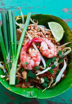 To zbliżenie talerza z danie Pad Thai, popularnym tajskim daniem z makaronem. Makaron jest smażony na wok z krewetkami, kiełkami fasoli, orzeszkami ziemnymi i szczypiorkiem. Danie podane jest na liściu banana, co jest typowe dla kuchni tajskiej. Jasnozielony i czerwony kolor potrawy są intensywne i apetyczne. Tło stanowi rustykalny drewniany stół pomalowany na turkusowo.