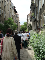 Uliczka między kamienicami, a na niej spacerująca grupa osób.