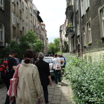 Uliczka między kamienicami, a na niej spacerująca grupa osób.