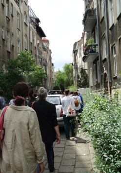 Uliczka między kamienicami, a na niej spacerująca grupa osób.