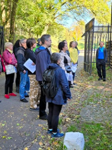 Obraz przedstawia grupę ludzi stojących przed ceglanym murem, słuchających mężczyzny, który wskazuje na coś na ścianie. Za nimi znajdują się drzewa, a w tle brama. To jasny słoneczny dzień.