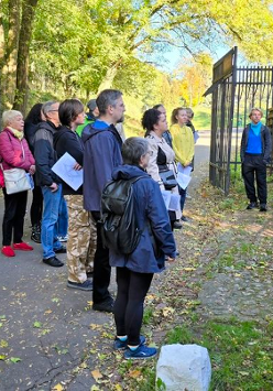 Obraz przedstawia grupę ludzi stojących przed ceglanym murem, słuchających mężczyzny, który wskazuje na coś na ścianie. Za nimi znajdują się drzewa, a w tle brama. To jasny słoneczny dzień.