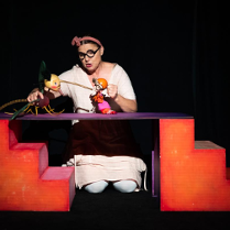 Zdjęcie ze spektaklu - aktorka klęcząc za elementem scenografii trzyma w rękach dwie lalki.