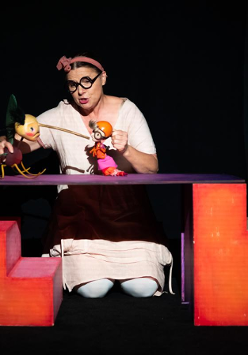 Zdjęcie ze spektaklu - aktorka klęcząc za elementem scenografii trzyma w rękach dwie lalki.