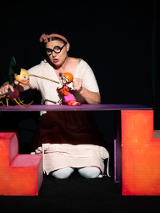 Zdjęcie ze spektaklu - aktorka klęcząc za elementem scenografii trzyma w rękach dwie lalki.