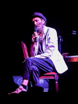 Zdjęcie ze spektaklu: przy pianinie, odwrócony do niego tyłem, na krześle siedzi mężczyzna z mikrofonem w ręce.