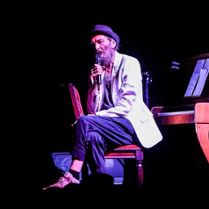 Zdjęcie ze spektaklu: przy pianinie, odwrócony do niego tyłem, na krześle siedzi mężczyzna z mikrofonem w ręce.