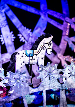 Zdjęcie ze spektaklu - biały drewniany konik wśród dekoracji w bialo-fioletowych kolorach.