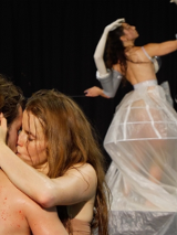 Zdjęcie ze spektaklu. Na pierwszym planie całująca się para. W tle kobieta w bialej przezroczystej sukni.