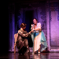 Zdjęcie ze spektaklu - dwie kobiety na scenie, jedna siedzi na krześle, druga stoi i trzyma w rękach lalkę.
