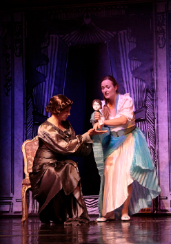 Zdjęcie ze spektaklu - dwie kobiety na scenie, jedna siedzi na krześle, druga stoi i trzyma w rękach lalkę.