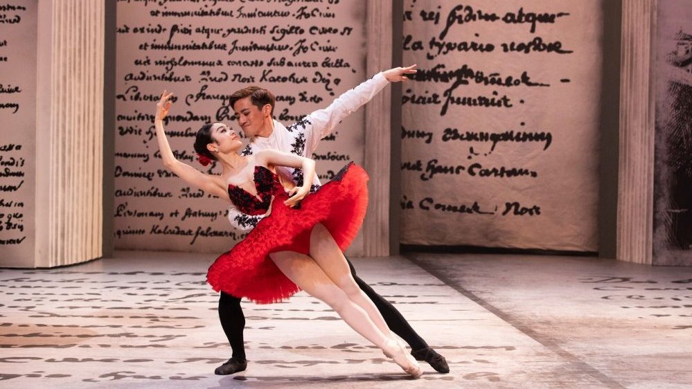 Para tancerzy baletowych wykonuje figurę na tle scenografii z motywem zapisanych starych ksiąg.