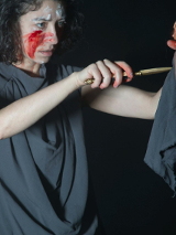 Zdjęcie ze spektaklu - kobieta z zakrwawioną twarzą w jednej ręce trzyma lalkę, a w drugiej sztylet, który przykłada do lalki.