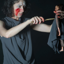 Zdjęcie ze spektaklu - kobieta z zakrwawioną twarzą w jednej ręce trzyma lalkę, a w drugiej sztylet, który przykłada do lalki.