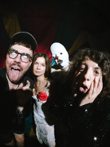 Czworo ludzi pozuje w teatralny sposób: dwie osoby robią śmieszne miny, jedna stoi poważnie, a czwarta w przebraniu cyrkowym. Scena ma cyrkowy klimat.