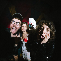 Czworo ludzi pozuje w teatralny sposób: dwie osoby robią śmieszne miny, jedna stoi poważnie, a czwarta w przebraniu cyrkowym. Scena ma cyrkowy klimat.