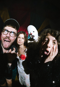 Czworo ludzi pozuje w teatralny sposób: dwie osoby robią śmieszne miny, jedna stoi poważnie, a czwarta w przebraniu cyrkowym. Scena ma cyrkowy klimat.