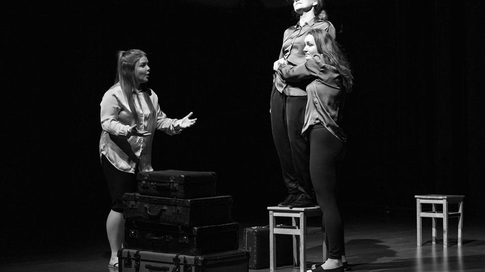Czarno-białe zdjęcie. Trzy kobiety na scenie. Jedna stoi na krześle z zamknietymi oczami. Druga przytula się niej. Trzecia mówi coś, zwracając sie do pozostałych, gestykuluje.
