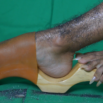 Zdjęcie owłosionej nogi pokrytej brokatem w drewnianym chodaku. Obok fragment drugiej stopy z długimi paznokciami.