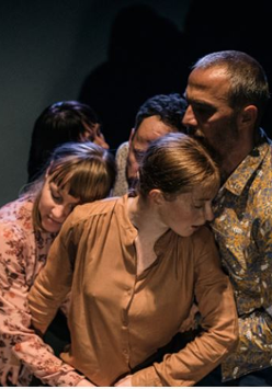 Zdjęcie przedstawia grupę aktorów w trakcie trwania spektaklu. Po lewej stronie znajdują się trzy kobiety, po prawej dwóch mężczyzn. W tle widoczne są dwie nieoświetlone sylwetki, sprawiające wrażenie kryjących się w cieniu.