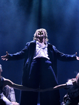 Zdjęcie ze spektaklu - główny bohater śpiewa na scenie w otoczeniu tancerzy.