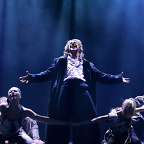 Zdjęcie ze spektaklu - główny bohater śpiewa na scenie w otoczeniu tancerzy.