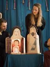 Zdjęcie ze spketaklu: trzech aktorów, jeden z nich gra na instrumencie przypominającym mandolinę, drugi trzyma drewnianą rzeźbę. Na stoliku pomiędzy dwoma aktorami stoją trzy drewniane rzeźby przedstawiające świętą rodzinę, za stolikiem stoi aktorka.