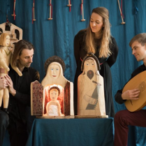 Zdjęcie ze spketaklu: trzech aktorów, jeden z nich gra na instrumencie przypominającym mandolinę, drugi trzyma drewnianą rzeźbę. Na stoliku pomiędzy dwoma aktorami stoją trzy drewniane rzeźby przedstawiające świętą rodzinę, za stolikiem stoi aktorka.