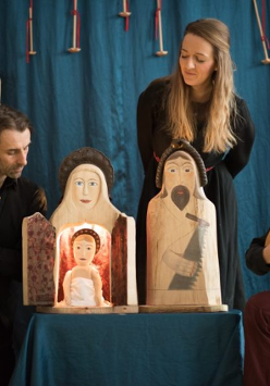 Zdjęcie ze spketaklu: trzech aktorów, jeden z nich gra na instrumencie przypominającym mandolinę, drugi trzyma drewnianą rzeźbę. Na stoliku pomiędzy dwoma aktorami stoją trzy drewniane rzeźby przedstawiające świętą rodzinę, za stolikiem stoi aktorka.