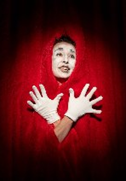 Postać w czerwonym stroju i wymalowaną na biało twarzą stoi na tle kurtyny teatralnej.