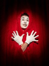 Postać w czerwonym stroju i wymalowaną na biało twarzą stoi na tle kurtyny teatralnej.