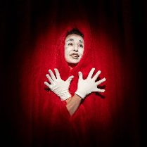 Postać w czerwonym stroju i wymalowaną na biało twarzą stoi na tle kurtyny teatralnej.