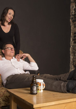 Zdjęcie ze spektaklu - kobieta stoi za siedzącym na fotelu mężczyźnie i opiera mu dłonie na ramianach. Mężczyzna ma położone nogi na stoliku, na którym stoi też butelka i kubek.