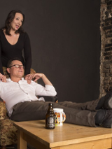 Zdjęcie ze spektaklu - kobieta stoi za siedzącym na fotelu mężczyźnie i opiera mu dłonie na ramianach. Mężczyzna ma położone nogi na stoliku, na którym stoi też butelka i kubek.