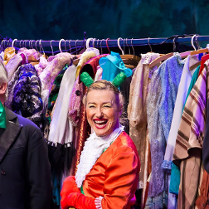 Zdjęcie ze spektaklu - aktorka w przebraniu do przedstawienia na tle wieszaka pełnego ubrań.